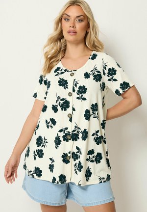 Mujer que lleva una camiseta blanca de manga corta con botones y estampado floral negro, y pantalones cortos vaqueros azul claro, de pie frente a un fondo liso.