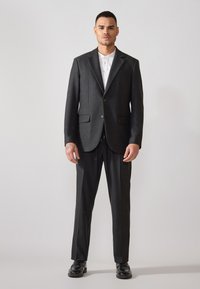Traje de corte ajustado en gris oscuro con chaqueta de un solo botón, solapas de muesca, bolsillos de solapa y pantalones a juego. Camisa blanca con cuello debajo.