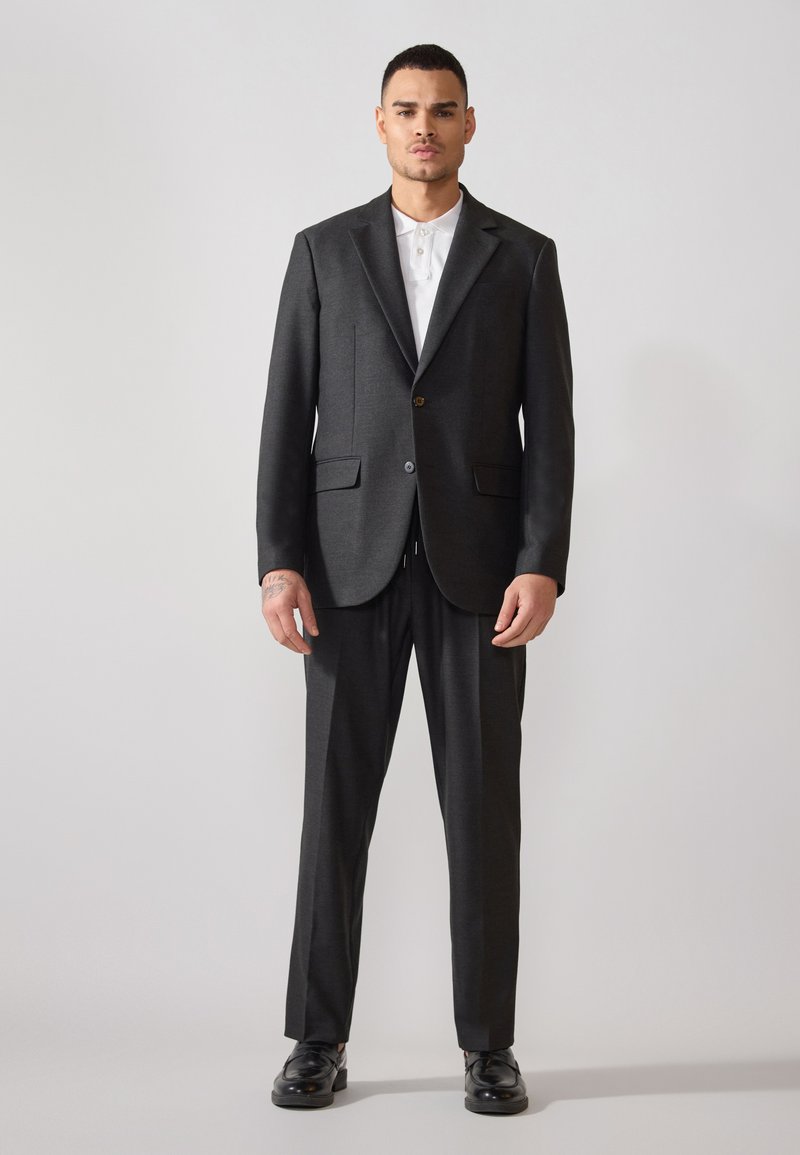 Traje de corte ajustado en gris oscuro con chaqueta de un solo botón, solapas de muesca, bolsillos de solapa y pantalones a juego. Camisa blanca con cuello debajo.