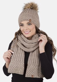 Berretto beige in maglia con pompon in pelliccia sintetica e sciarpa coordinata con motivi testurizzati e etichetta in pelle, indossato da una persona.