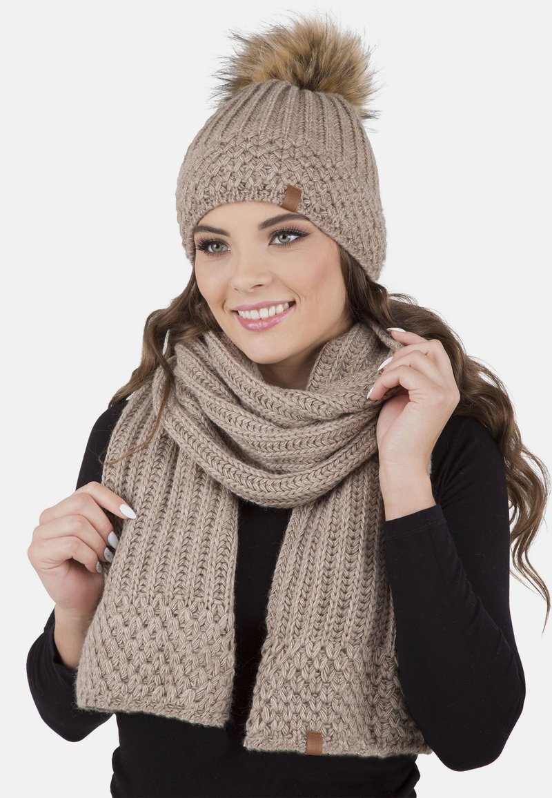 Berretto beige in maglia con pompon in pelliccia sintetica e sciarpa coordinata con motivi testurizzati e etichetta in pelle, indossato da una persona.