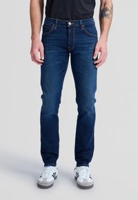 Homme portant un jean slim bleu foncé, un t-shirt noir, des baskets blanches avec des bandes noires et un tatouage sur son avant-bras gauche.