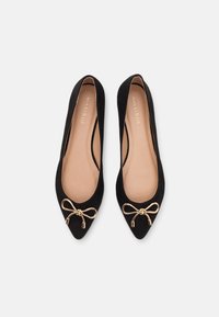 Ballerines en daim noir avec un bout pointu, dotées d'un accent nœud doré à l'avant et d'une semelle intérieure beige lisse.