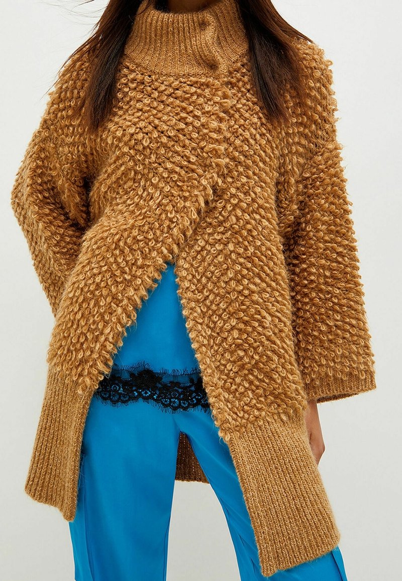 Flauschige Strickjacke in Braun mit hohem Kragen, ausgestattet mit strukturierten Bobbles. Kombiniert mit blauen Satin-Hosen und einem schwarzen Spitzenbesatz.