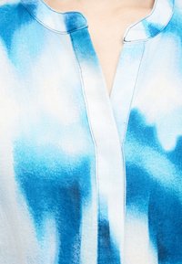 Εγγύς λήψη πουκάμισου χωρίς γιακά μπλε-άσπρου tie-dye με άνοιγμα σε σχήμα V σε ανοιχτό τόνο δέρματος.