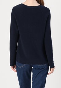 Personne portant un pull en maille bleu marine à manches longues et un jean bleu foncé, vue de dos sur un fond uni.