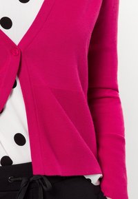 Cardigan fucsia in a maglia texturizzata con scollo a V, chiusura a un bottone e sottili dettagli a coste; indossato sopra una camicia a pois.