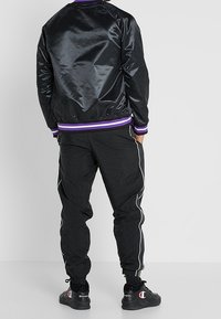 Chaqueta bomber de satén negra con puños y cuello de punto morado, combinada con pantalones jogger negros que tienen rayas laterales blancas y tobillos ajustados.