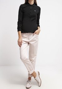 Polo noir à manches longues avec fermeture à boutons et logo, associé à un pantalon tailleur rose clair et des chaussures de ville claires.