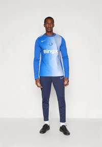 Maglia sportiva a maniche lunghe blu con design a gradiente, logo e testo sulla parte anteriore, abbinata a pantaloni della tuta blu navy e scarpe nere.