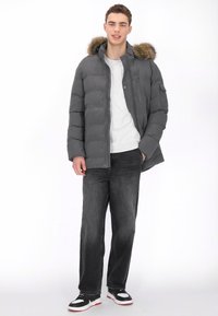 Kurtka puchowa w ciemnoszarym kolorze z kapturem wyściełanym futrem, zapinana na zamek i guziki, noszona na jasnoszarej koszuli i czarnych dżinsach.