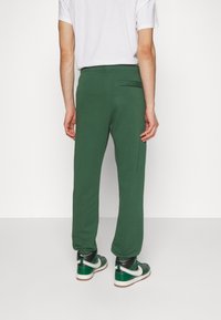 Man som bär gröna träningsoverallsbyxor, vit t-shirt och gröna, vita och svarta sneakers, stående mot en vit vägg med ryggen mot kameran.