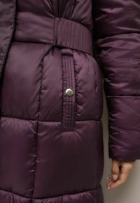 Nööritud lilla puffer-jakk, millel on vööga talje, hõbedased nupud ja sile, läikiv tekstuur horisontaalsete paneelidega.