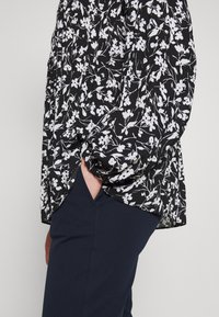 Blusa nera a fiori con motivi bianchi, caratterizzata da maniche arricciate e spacchi laterali. Abbinata a pantaloni blu navy, che mostrano una trama liscia.