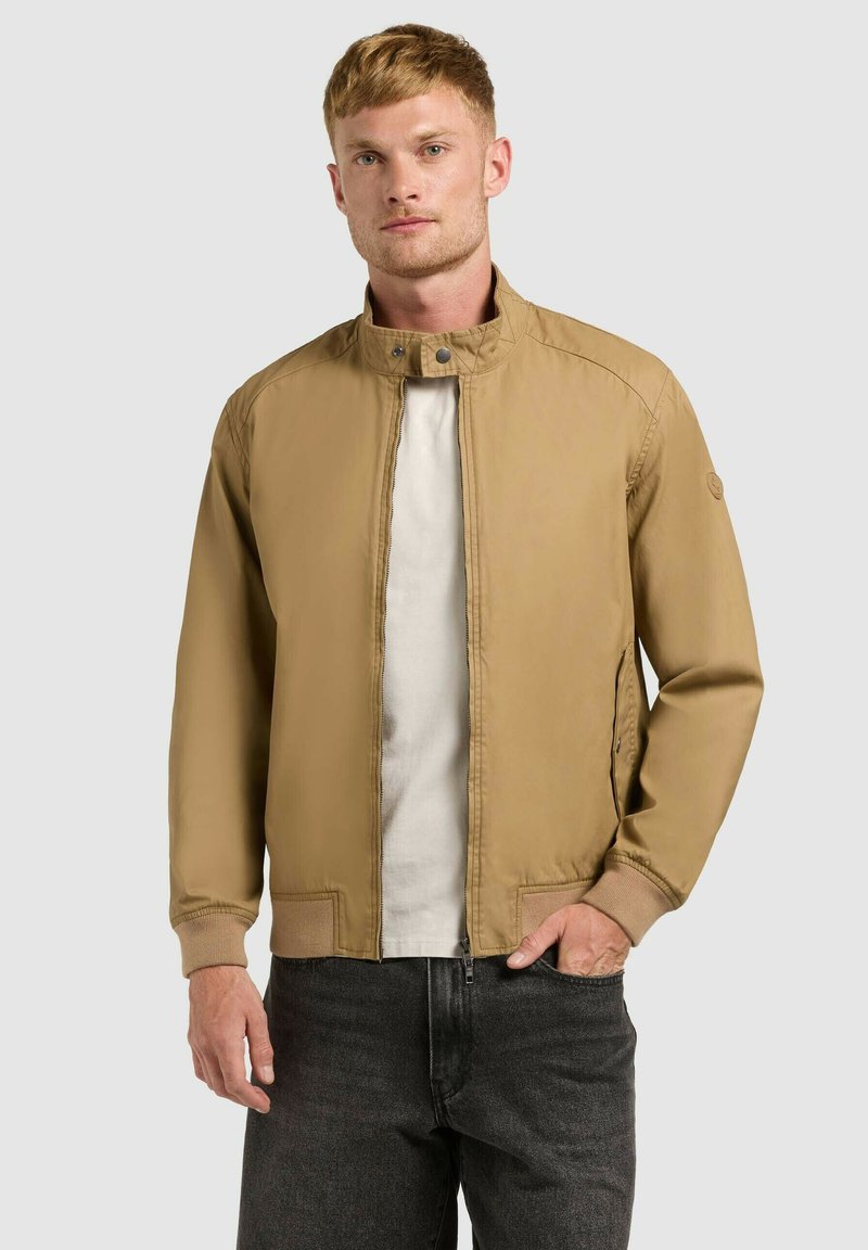 Homme portant une veste beige à fermeture éclair avec col montant, chemise blanche et jean noir, sur fond gris uni.