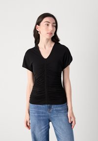 Black short-sleeve top with a V-neck, featuring front ruching details and a fitted shape. Paired with light blue denim jeans.