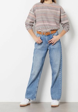 Vrouw die lichtblauwe high-waist jeans, een gebreide trui met patroon, een bruine riem en witte sneakers draagt, staand tegen een witte muur.