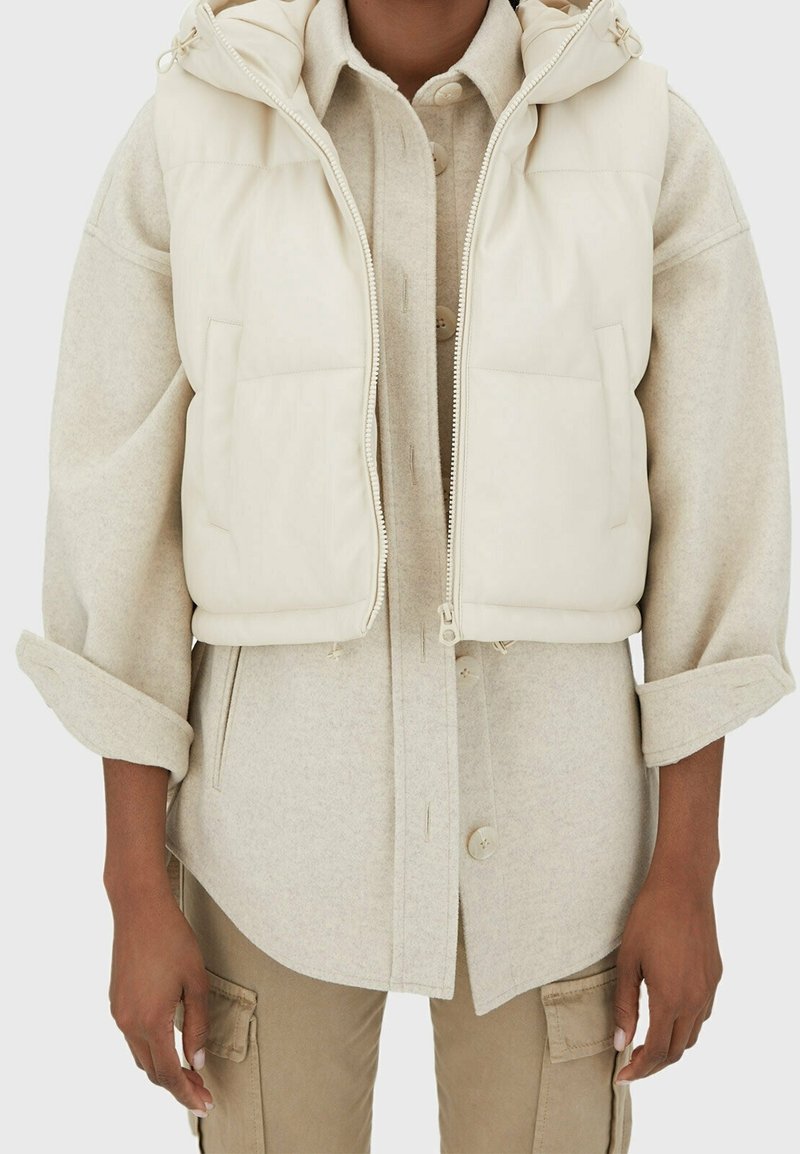 Personne portant une veste matelassée crème par-dessus une chemise beige à boutons avec les manches retroussées et un pantalon cargo beige.