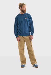 Felpa blu con collo a giro e logo ricamato, abbinata a pantaloni cargo beige con tasche laterali. Sneakers nere e blu completano il look.