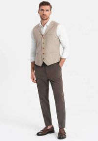 Chaleco de tejido beige con botones de madera, combinado con pantalones marrones rayados oscuros y zapatos marrones sin cordones, todo sobre un fondo liso.