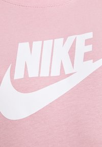 Růžové bavlněné tričko s velkým bílým nápisem "NIKE" a grafikou loga swoosh na přední straně, s hladkou texturou a kulatým výstřihem.