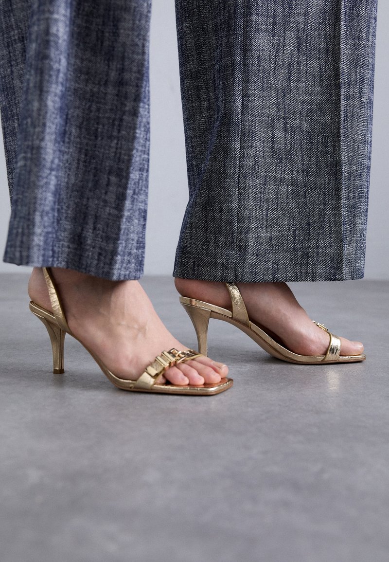 Sandales à talons hauts dorées au design élégant, avec une bride traversant les orteils et une hauteur de talon d'environ 7 cm. Fini métallique texturé.