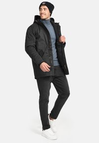 Chaqueta negra aislante con capucha y cierre de cremallera, llevada sobre un suéter gris. Combinada con jeans negros y zapatillas blancas. Fondo claro.