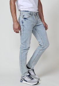 Muž oblečený v svetlomodrých slim-fit džínoch a sivých teniskách stojí pred jednoliatym pozadím.
