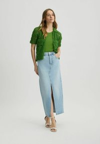 Green crochet cardigan over a green tank top, paired with a light blue denim maxi skirt featuring a front slit. White strappy heels.