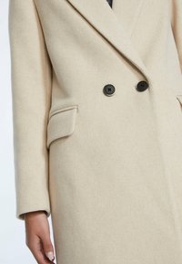 Primer plano de un abrigo beige de doble botonadura con botones negros, bolsillos de solapa y la mano de una persona con uñas de color claro descansando junto a la manga.
