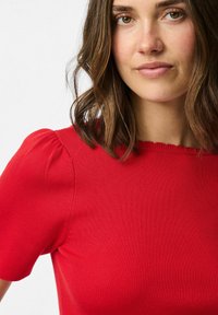 Top rosso a coste con maniche a sbuffo corte, caratterizzato da un collo ondulato. Il tessuto appare texturizzato e liscio.