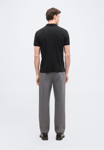 Homme debout de dos, portant un polo noir à manches courtes, un pantalon gris et des chaussures habillées noires, devant un fond uni.