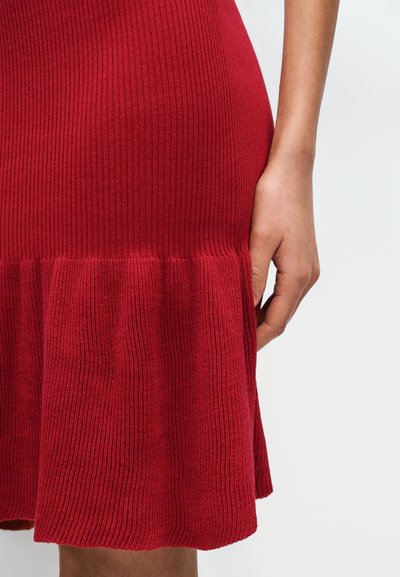 Κόκκινο ζιβάγκο φόρεμα με ριμπ ύφανση και φαρδιά τελείωμα, με υφή υφάσματος και εφαρμοστό μπούστο, με το χέρι τοποθετημένο στη μέση στο πλάι.