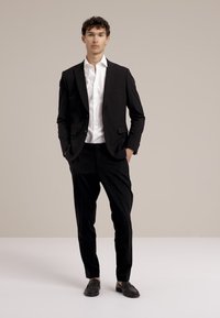 Črn kostim s slim krojem, bela srajca in črne loafer čevlje. Gladek material, prilegajoč kroj in ravne hlače. Preprost, minimalističen stil.
