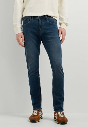 Homme portant un jean slim bleu foncé, un pull crème et des baskets marron avec des bandes blanches, debout devant un fond uni.
