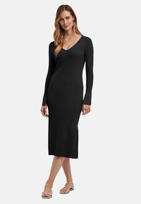Abito a maglia nero a coste con maniche lunghe e scollo a V profondo. Presenta una vestibilità aderente e lunghezza fino al polpaccio. Indossato con sandali con cinturini.