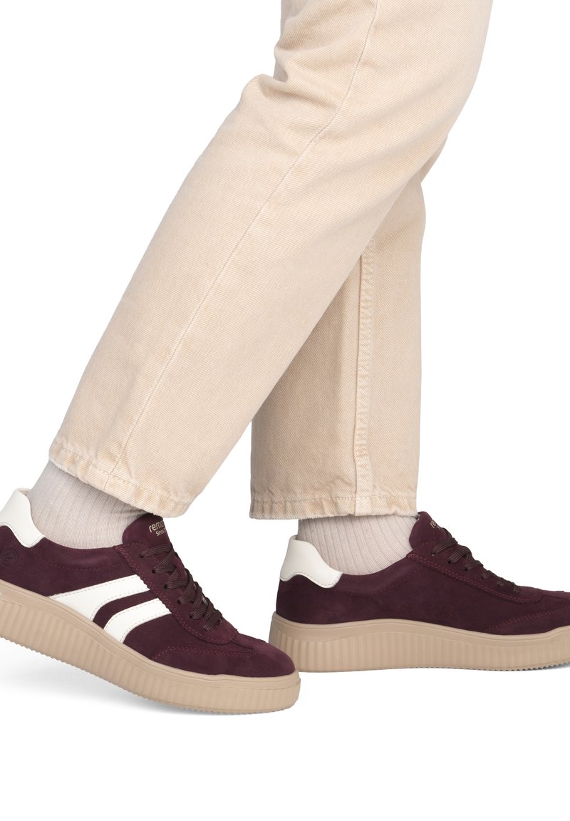 Personne portant un pantalon beige, des chaussettes blanches et des baskets bordeaux à bandes blanches et semelles beiges, avançant sur un fond uni.