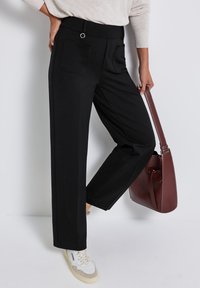 Pantalons larges noirs avec une texture lisse, dotés de deux poches arrière, d'une taille plate et d'un accent en métal circulaire. Assortis avec un sac à main marron.