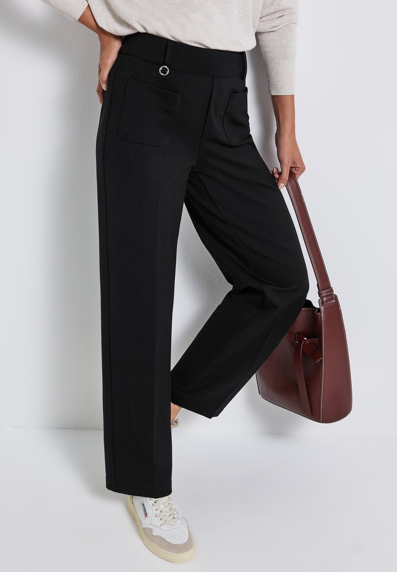 Pantalons larges noirs avec une texture lisse, dotés de deux poches arrière, d'une taille plate et d'un accent en métal circulaire. Assortis avec un sac à main marron.