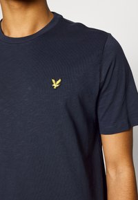 T-shirt blu navy realizzato in morbido cotone con scollatura rotonda e maniche corte. Presenta un piccolo logo di un'aquila ricamata di colore giallo sul petto.
