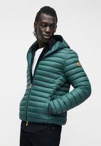 Άνδρας που φοράει τυρκουάζ μπουφάν με κουκούλα και επένδυση πάνω από μαύρο πουκάμισο και λευκό μπλουζάκι, κοιτάζοντας προς τα αριστερά του με τα χέρια στις τσέπες του μπουφάν.