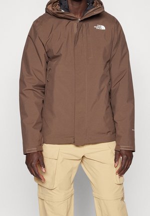 Mężczyzna ubrany w brązową kurtkę z kapturem zapinaną na zamek z przodu oraz beżowe spodnie typu cargo, stojący z rękami luźno opartymi wzdłuż ciała.