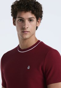 Bordeaux kortgemouwen gebreide shirt met een gestructureerd patroon, contrasterende witte kraagafwerking en klein geborduurd pinguïnlogo op de borst.