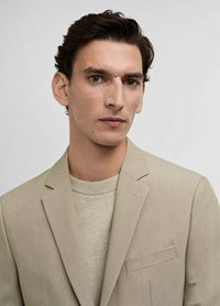 Beige blazer med strukturerad tyg, en bröstficka och slagna rever. Bärs över en ljusbeige tröja med rund halsringning.