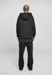 Rövid hajú személy háttal áll, fekete kapucnis pulóvert, sötét réteges inget, laza fekete nadrágot és fekete cipőt visel egyszínű háttér előtt.