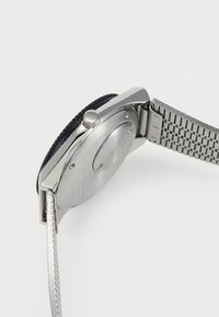 Montre-bracelet en métal argenté avec lunette noire, couronne visible et bracelet à maillons en métal, vue de côté en mettant l'accent sur l'arrière et la zone de la boucle.
