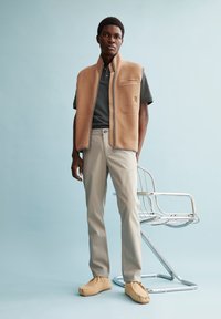 Gilet di pile color crema con colletto alto, tasche laterali e rifiniture grigie; abbinato a un polo grigia, pantaloni beige e scarpe slip-on color crema. Sedia in cromo sullo sfondo.