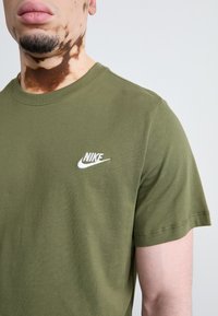 Olive spalvos „Nike“ marškinėliai, pagaminti iš medvilnės, su apvalia iškirptė ir trumpomis rankovėmis. Baltas „Nike“ logotipas išsiuvinėtas ant krūtinės.