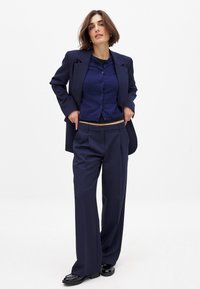 Navy-blau angezogener Anzug mit einem langen Sakko und weit geschnittenen Hosen, drübergezogen über einem enganliegenden navy-blauen Cardigan. Glattes Material und sichtbare Knopfdetails.