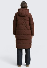 Braune Pufferjacke mit Kapuze, die über horizontale Steppnähte und ein knielanges Design verfügt. Kombiniert mit ausgestellten Jeanshosen.